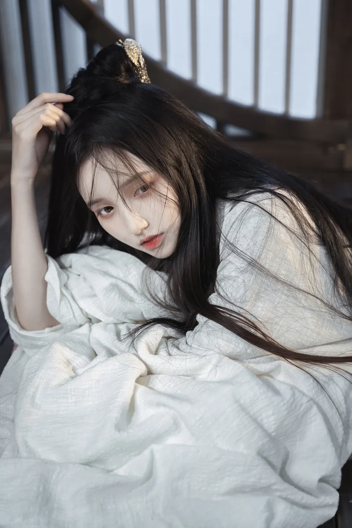 一名长发女子身穿白色衣服，侧身靠在室内柔软织物上，头发上饰有金色发饰。