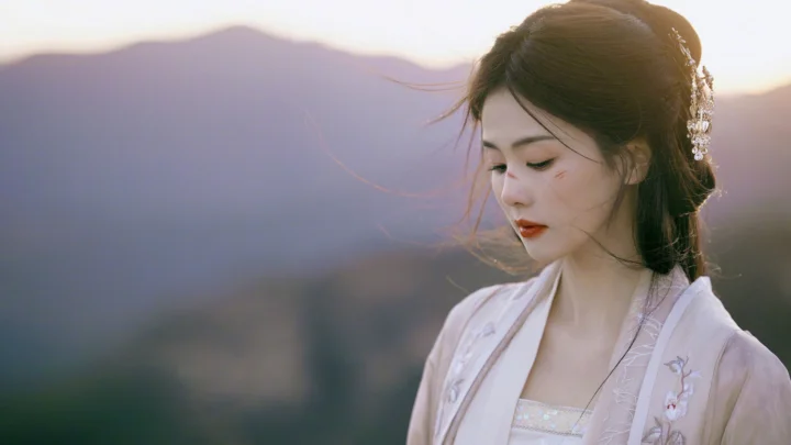 一名身穿浅色传统服饰的女子站在山景前，侧首低眸，阳光从后方照来。