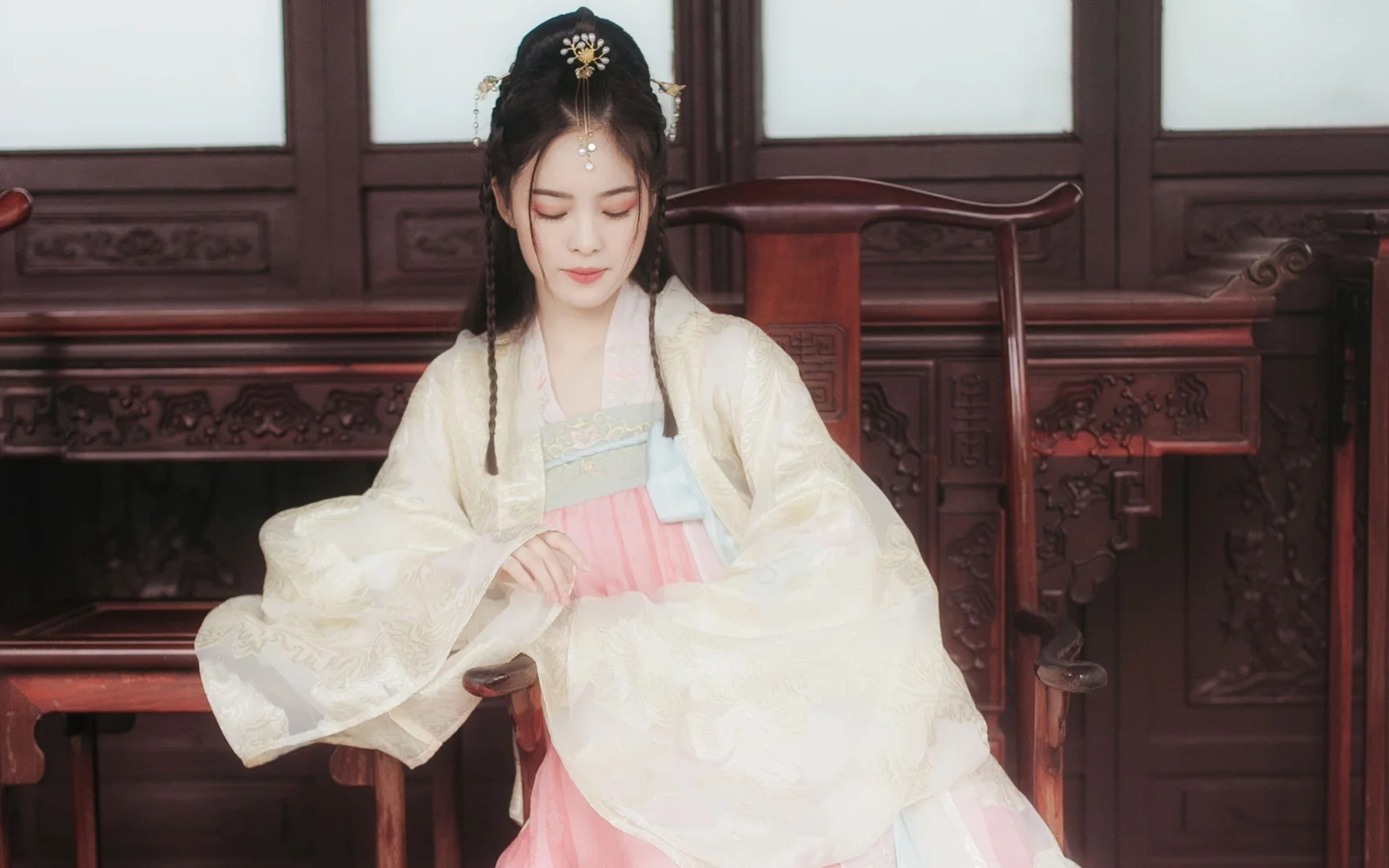 연한 색상의 한푸를 입은 여성이 조각된 목재 가구 앞 실내에 앉아 있다.