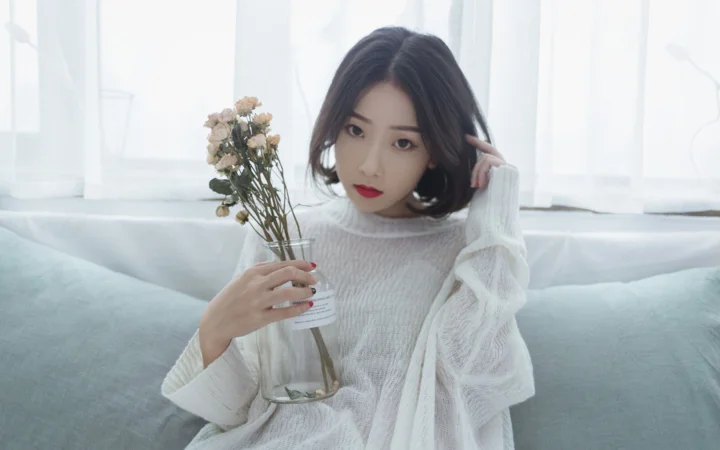 一名成年女性坐在明亮的室内沙发上，手持一束干花并望向镜头。