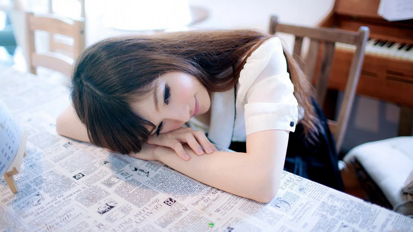 一名女子趴在铺满报纸的桌面上，背景可见室内钢琴。