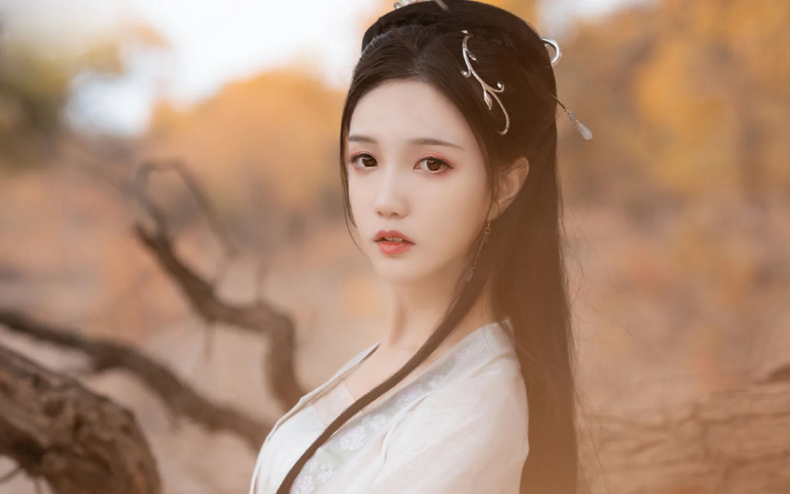 一名佩戴发饰、身穿白色传统服饰的女子站在秋色树木前。