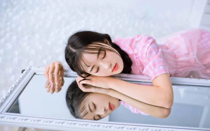 분홍색 체크 셔츠를 입은 여성이 거울 옆에 기대어 있고 거울에 모습이 비치고 있다.