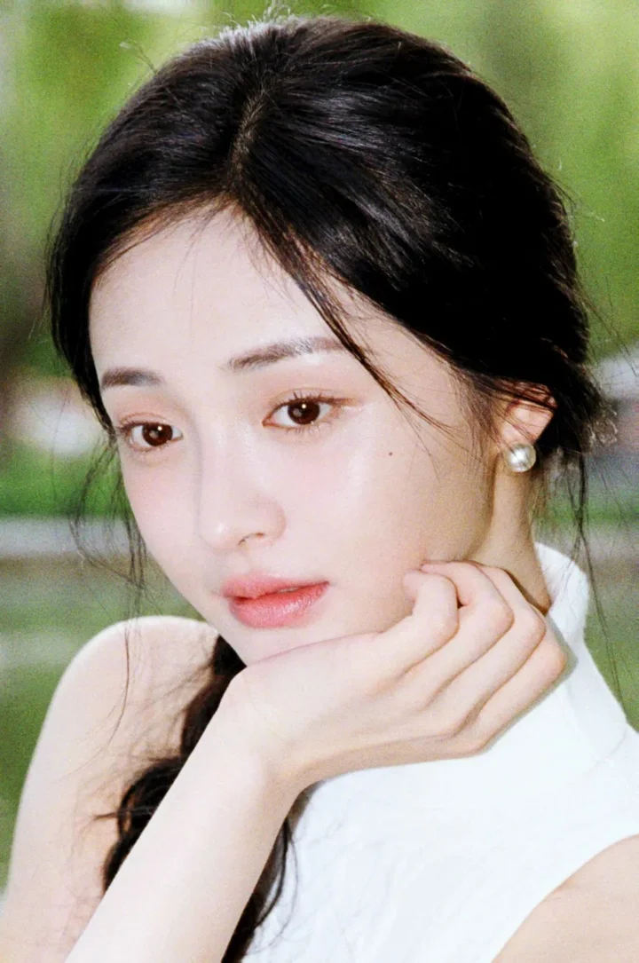 검은 머리 여성이 손으로 턱을 받치고 있는 클로즈업 인물 사진으로, 배경은 부드러운 녹색 흐림으로 보입니다.