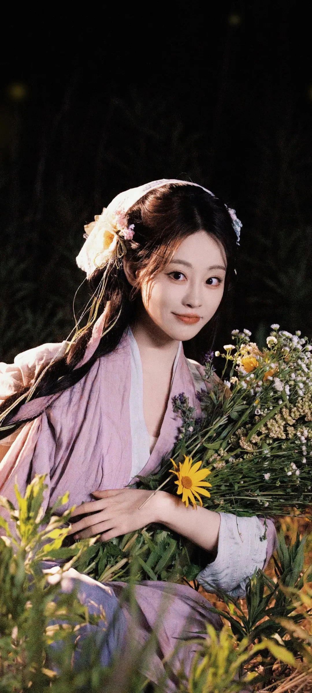 연보라색 옷을 입은 여성이 어두운 배경 앞에서 꽃다발을 안고 있다.