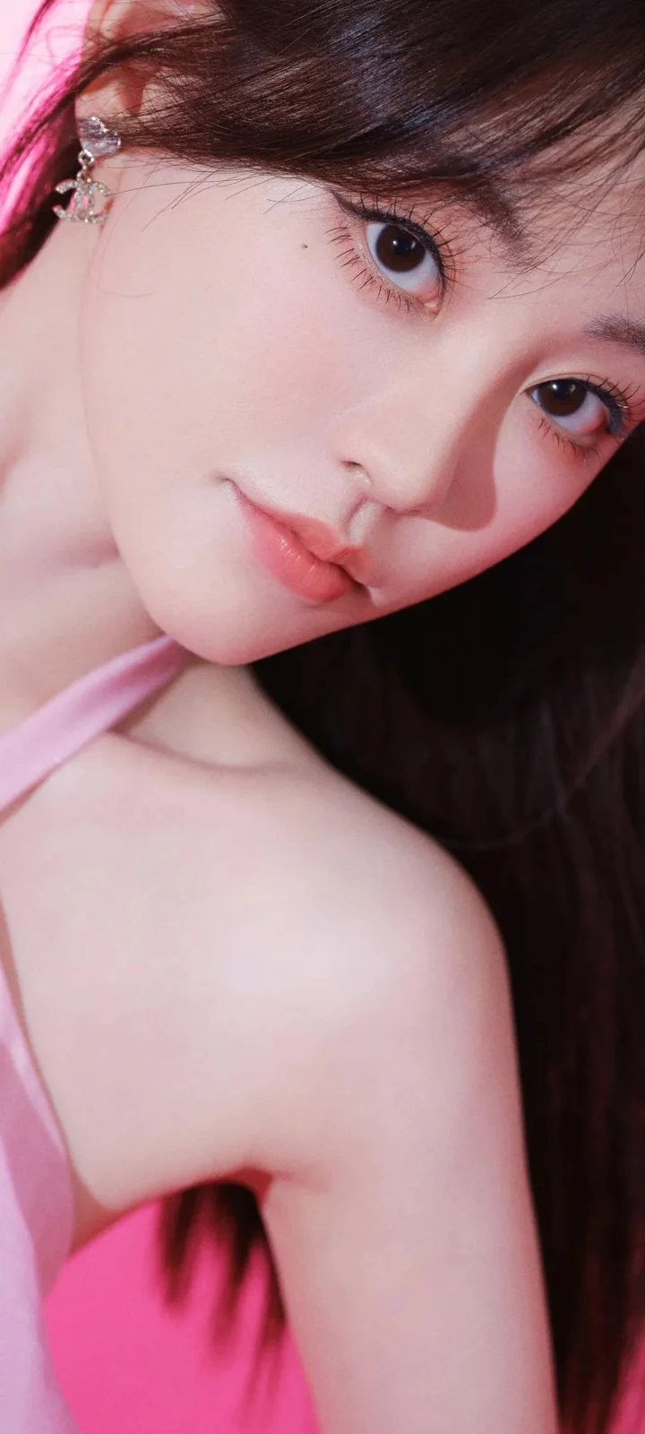一位深色长发女性以近距离姿态面对镜头，呈现粉色妆容和粉色衣带，背景较暗。