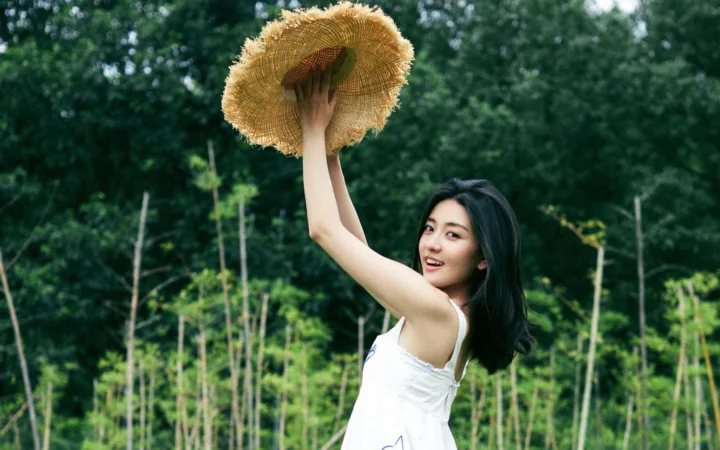 一名女子在绿色森林中举起草帽并面向镜头微笑。