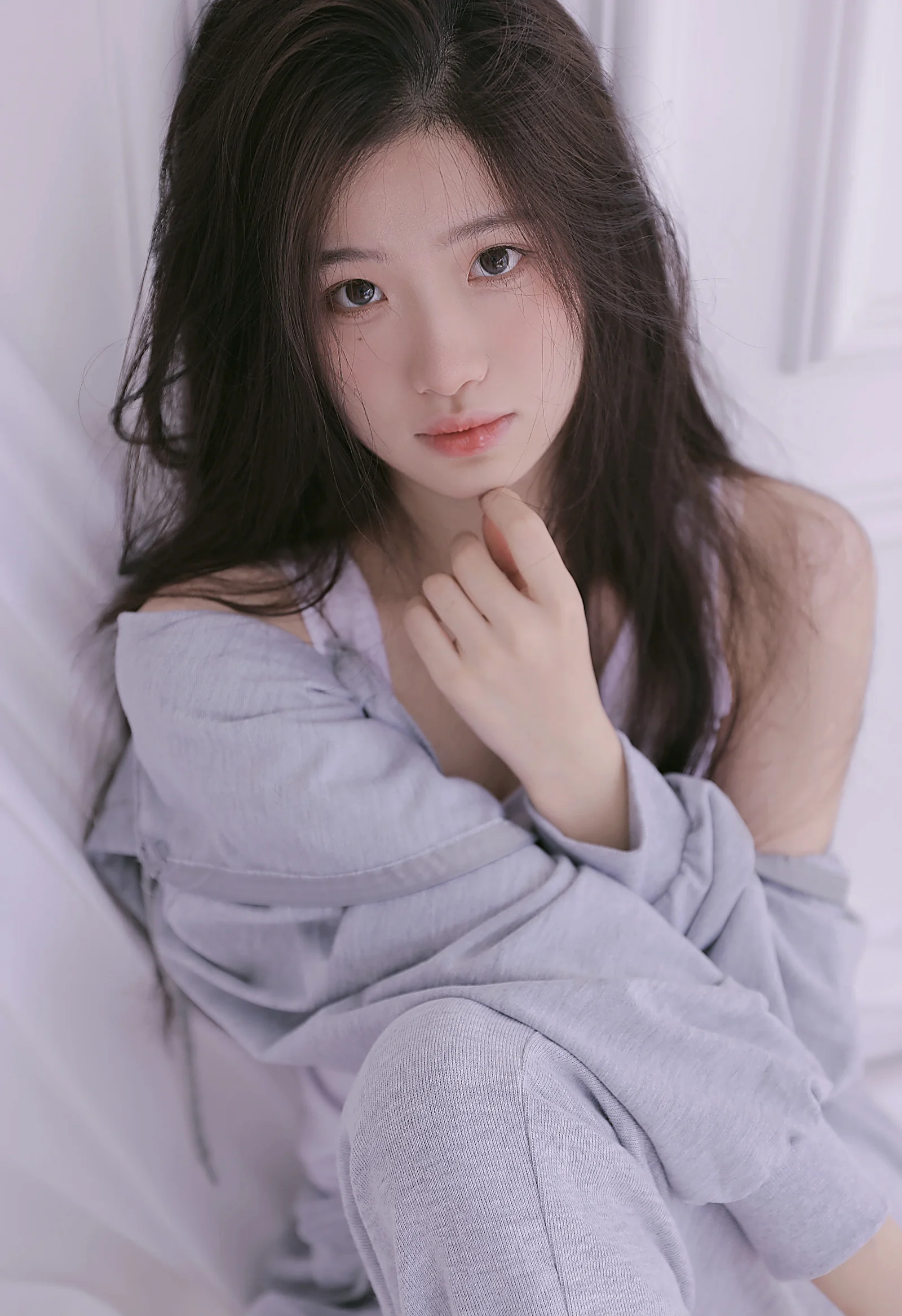 一名长发女性在柔和的淡紫色室内背景前抱膝坐着，手指轻触下巴并看向镜头。