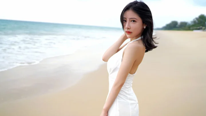 一名穿白色连衣裙的女子站在海滩上，身后是海水与沙滩。