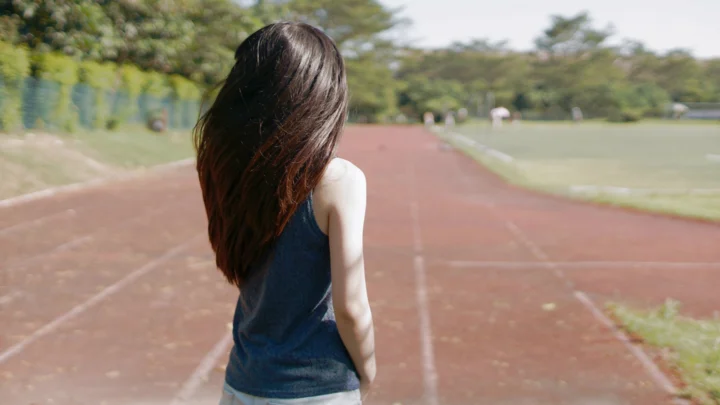 長い髪の女性が屋外の陸上トラックに背を向けて立っている。
