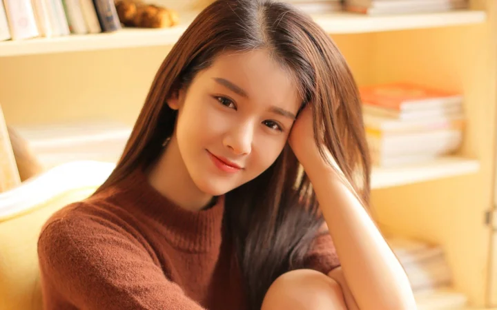 갈색 긴 머리의 여성이 갈색 니트 스웨터를 입고 따뜻하고 밝은 실내에서 팔에 기대어 카메라를 바라보고 있습니다.