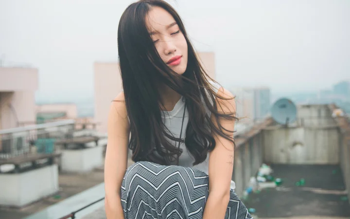 一名长发女子坐在城市屋顶上，远处可见模糊的建筑景观。