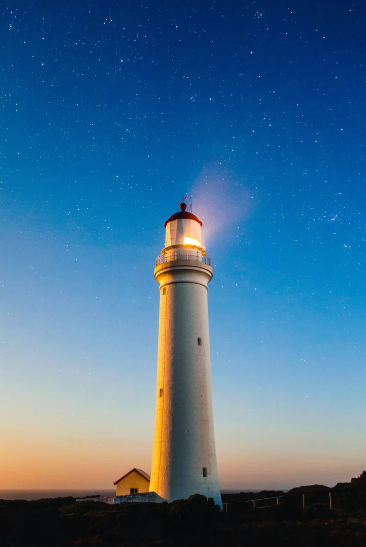 青い星空と夕暮れが広がる海岸に、白い灯台が立っています。