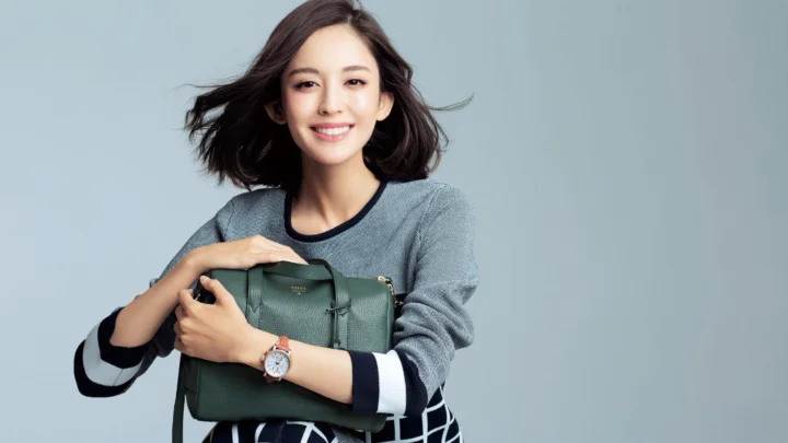 연한 회색 배경 앞에서 성인 여성이 짙은 초록색 핸드백을 안고 미소 짓고 있다.