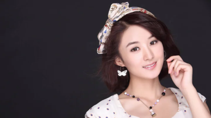 리본 헤어밴드와 귀걸이를 착용한 여성이 어두운 배경 앞에서 미소 짓고 있다.