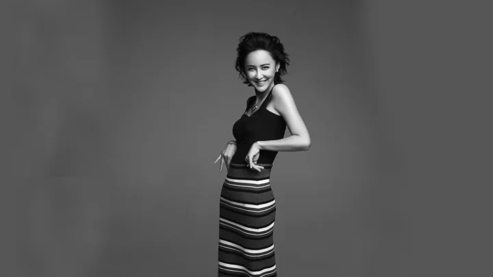 灰色背景前，一名微笑的成年女性身穿黑色上衣和条纹长裙摆姿势。