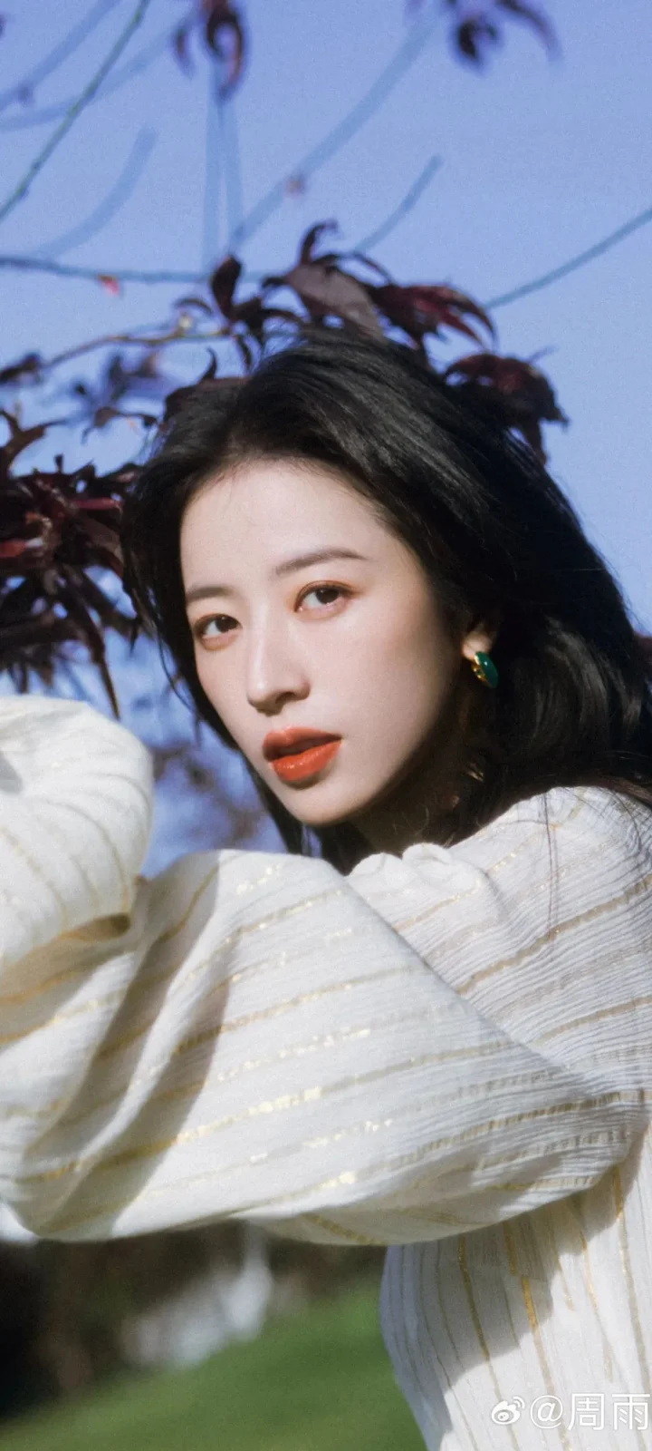 一名成年女性穿着白色毛衣，在户外树木背景前面向镜头。