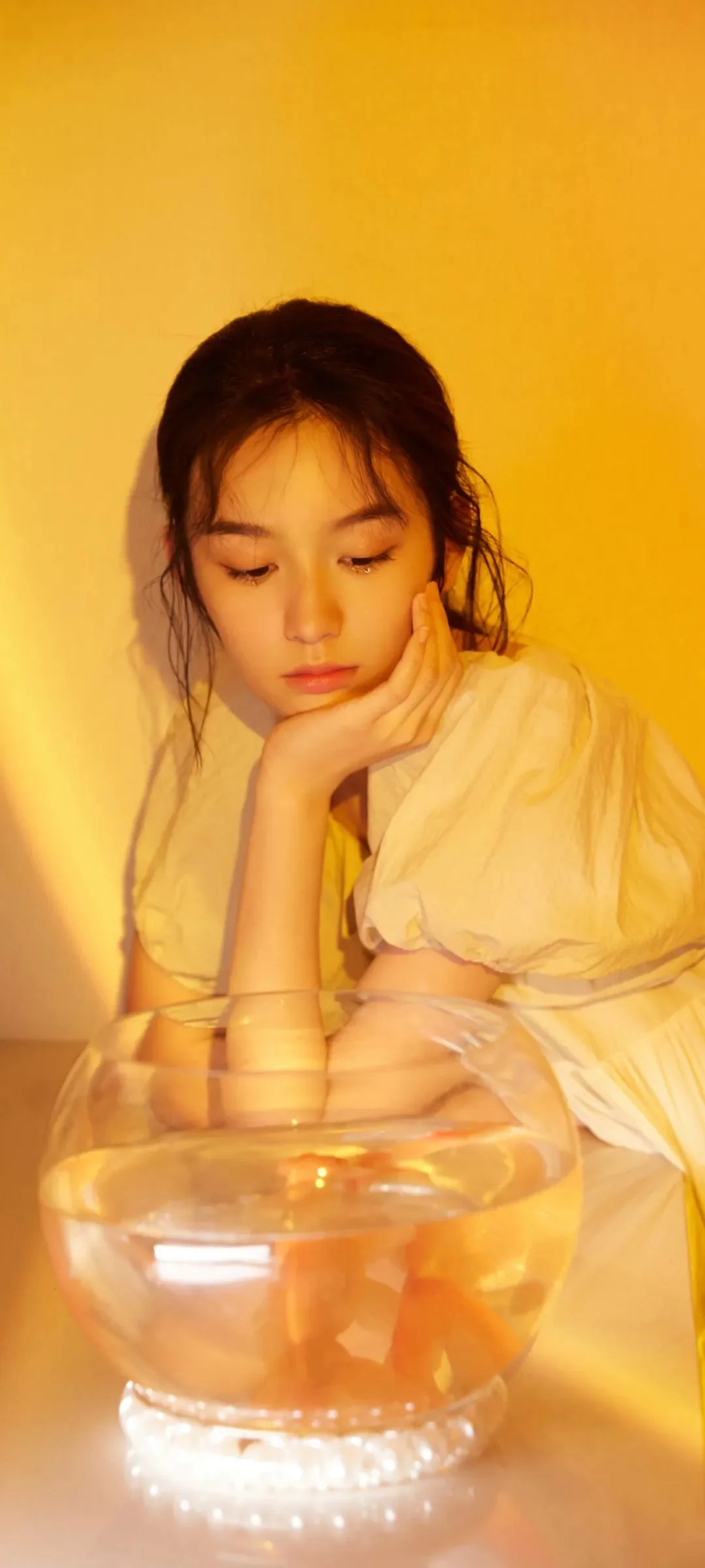 一名女性在温暖的黄色光线下托腮凝视前方的玻璃鱼缸。