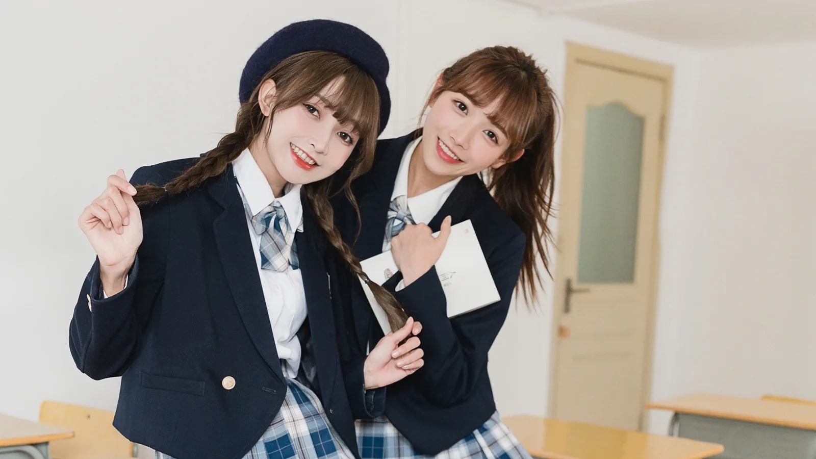 濃い色の制服を着た二人の女性が、室内で笑顔で並び、背景にドアと机が見えます。