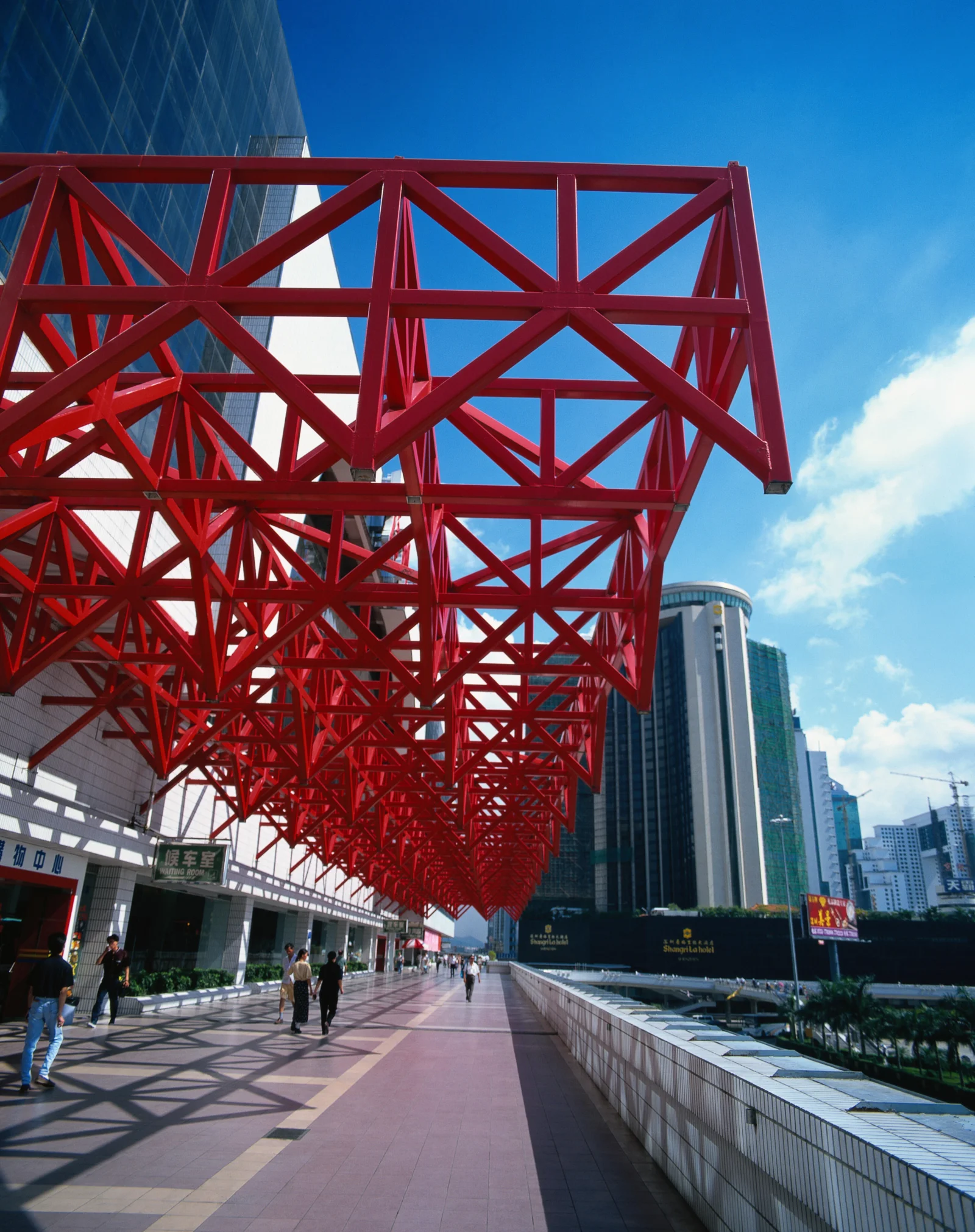 红色钢架结构横跨城市步道上方，周围可见现代高楼、蓝天和白云。