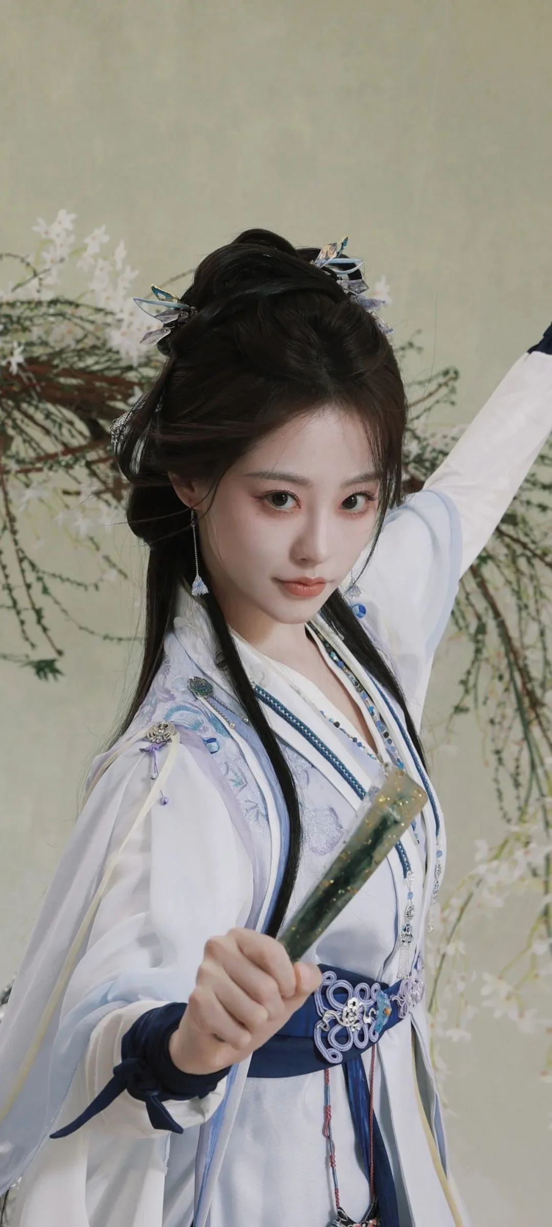 흰색 전통 의상을 입은 여성이 꽃가지를 들고 꽃 배경 앞에 서 있다.