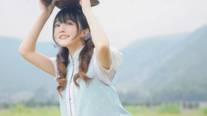 一位戴帽子的长发女子在山景与田野前微笑举帽。