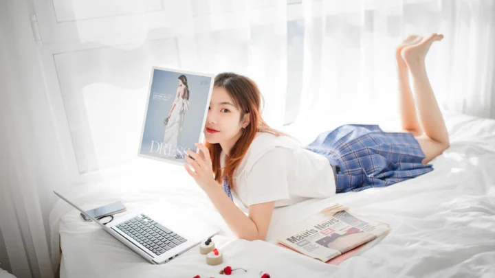 一名女性趴在白色床铺上，身旁放着笔记本电脑和杂志，手中拿着一本时尚册子。