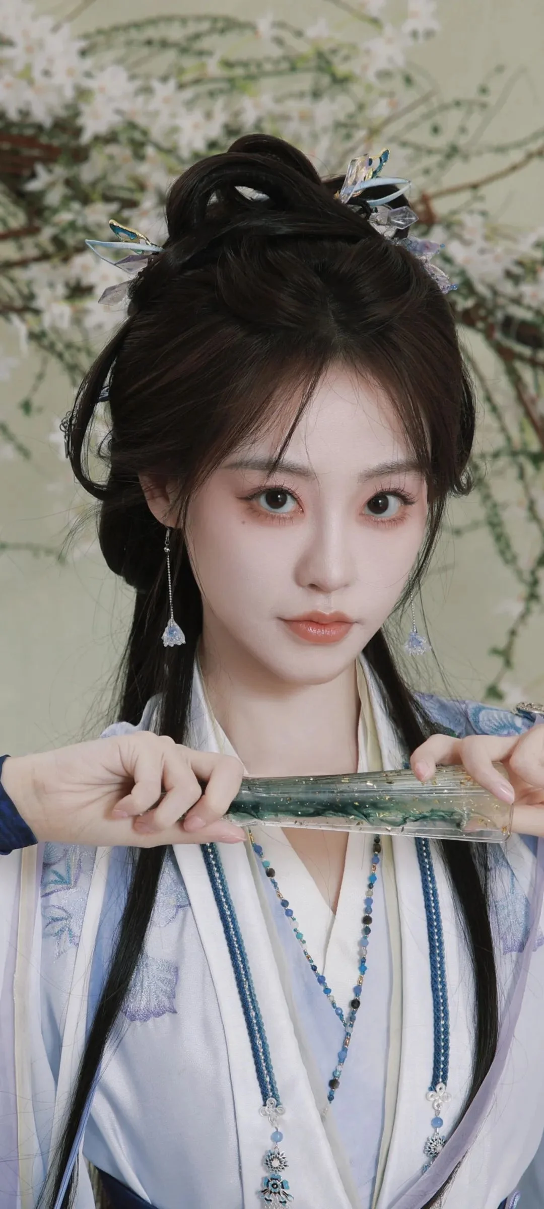 전통 의상을 입은 여성이 가느다란 물건을 들고 꽃무늬 배경 앞에 서 있다.