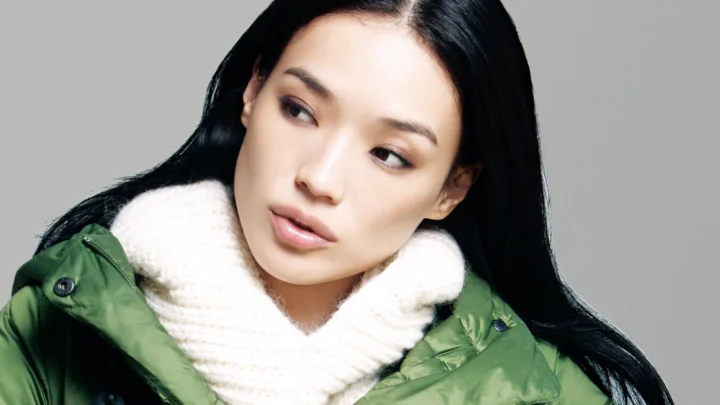 검은 머리의 여성이 초록색 겨울 재킷과 흰색 목도리를 두르고 회색 배경 앞에서 옆을 바라보고 있다.