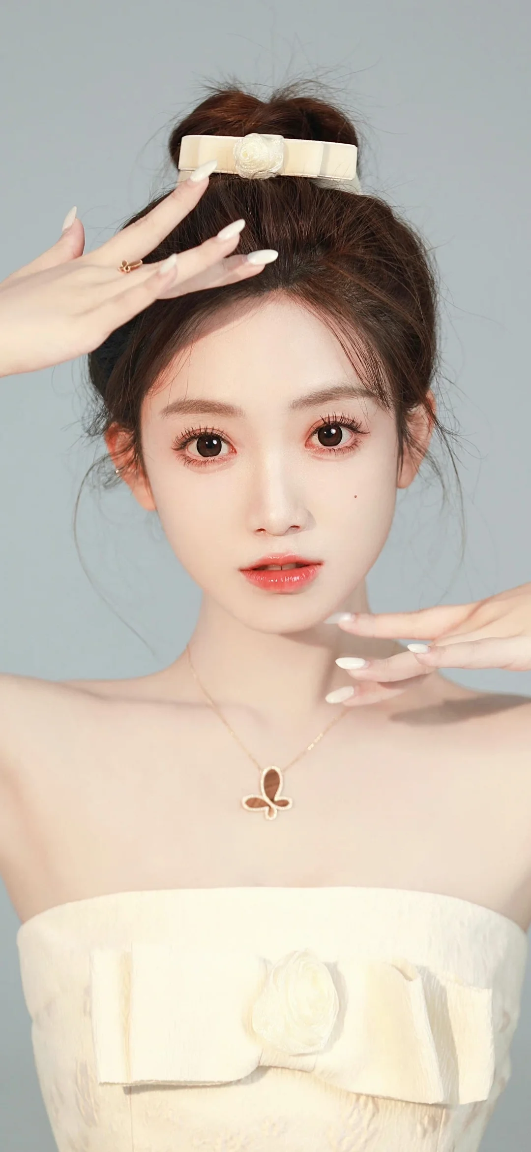 一位盘发女性身穿白色礼服，在浅灰色背景前直视镜头，双手靠近脸部。