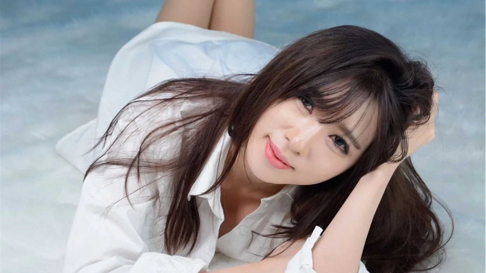긴 검은 머리의 성인 여성이 흰 셔츠를 입고 밝은 색 표면에 누워 있습니다.