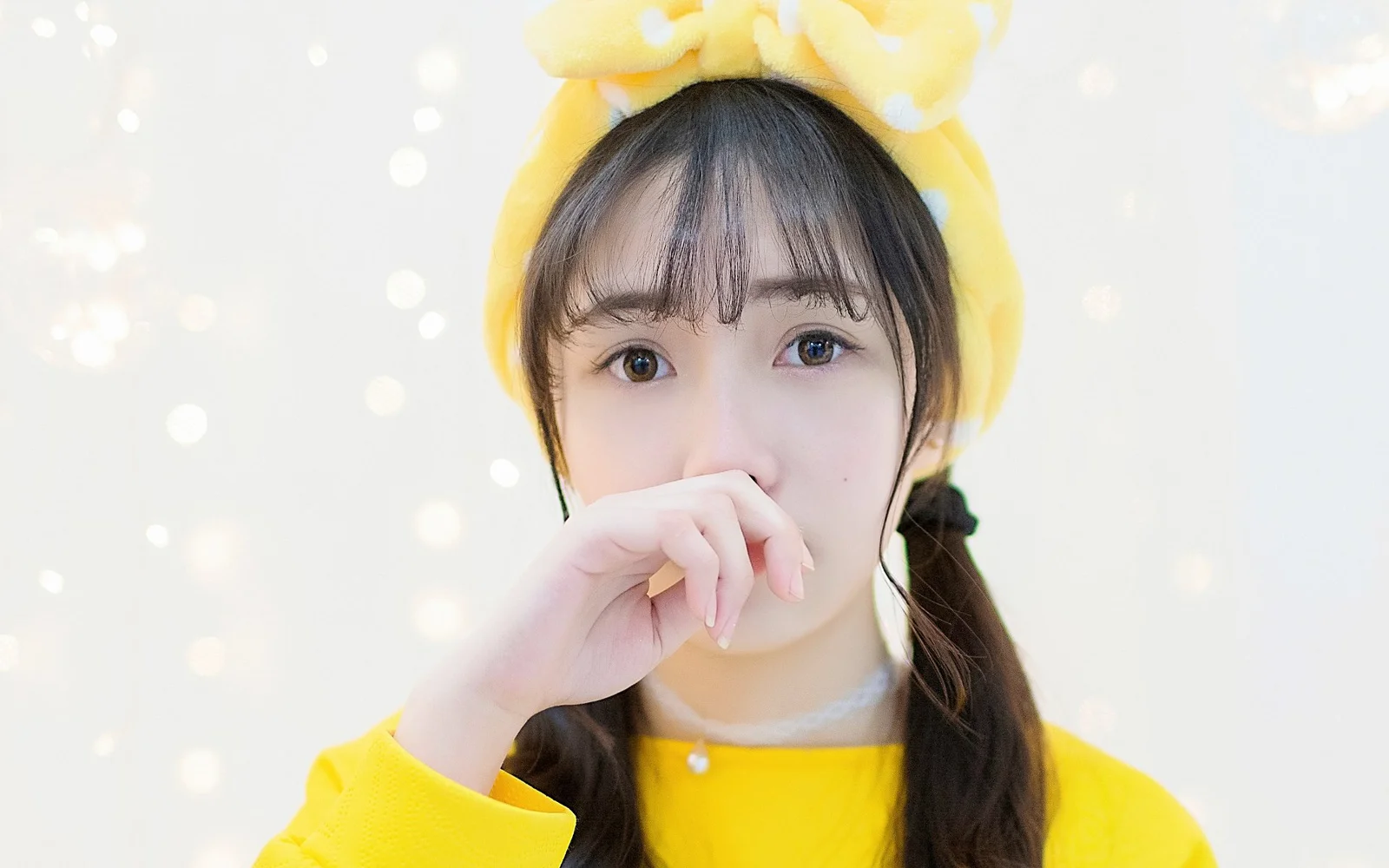 노란색 머리띠와 노란색 상의를 착용한 여성이 밝은 보케 배경 앞에서 손으로 입을 가리고 있습니다.