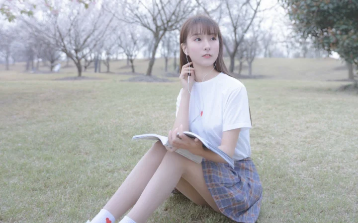 흰색 상의를 입은 여성이 공원 잔디에 앉아 책을 들고 있다.