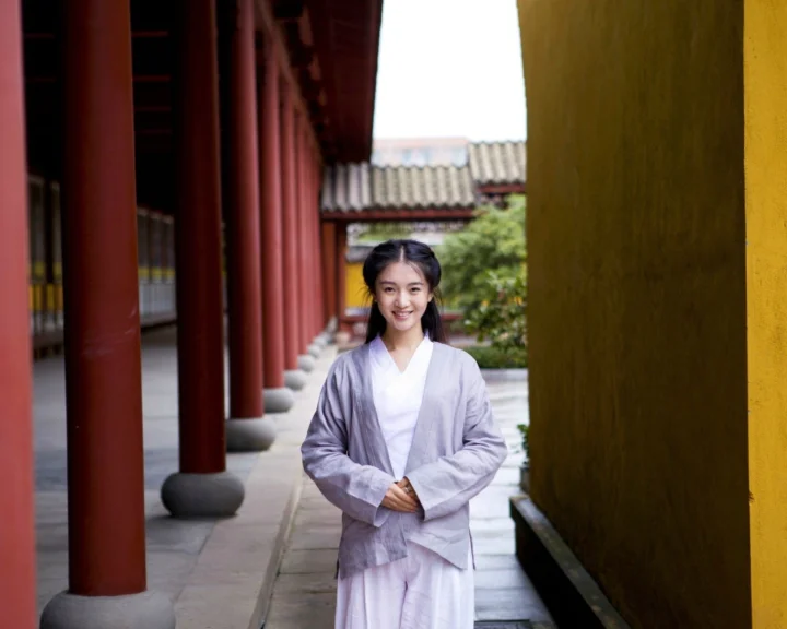 一名身穿浅色传统服饰的女子站在红柱与黄色墙面的庭院走廊中。