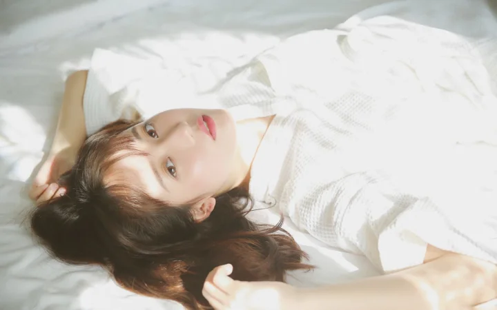 성인 여성이 침대에 누워 있으며 흰색 옷과 얼굴 위로 햇빛이 비치고 있습니다.