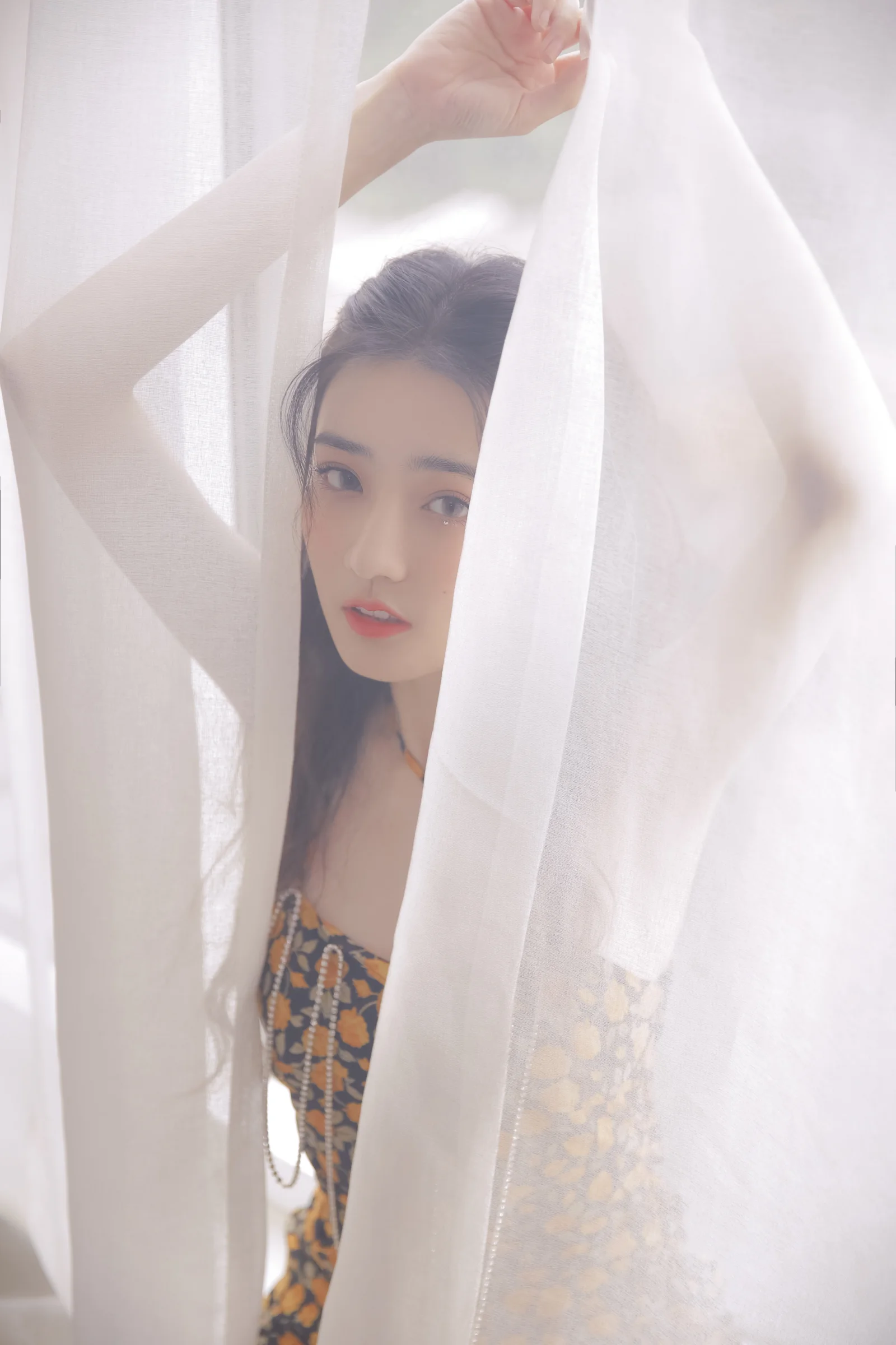 밝은 흰색 시어 커튼 뒤에서 여성이 카메라를 바라보고 있다.