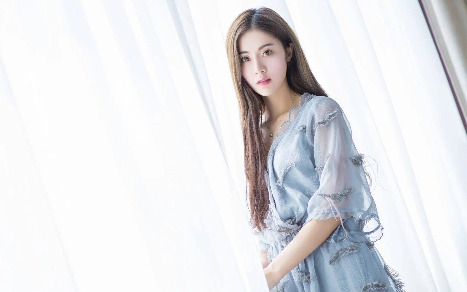 연한 파란색 옷을 입은 성인 여성이 흰 커튼 옆에 서 있습니다.