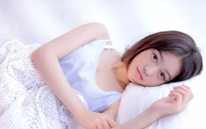 一名身穿白色上衣的女子侧躺在白色床铺上，面向镜头。