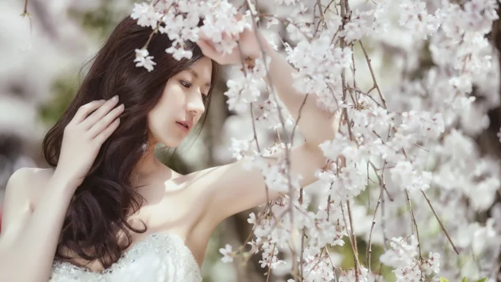 一位成年女子站在盛开的樱花枝旁，柔和光线映照出优雅的户外人像。