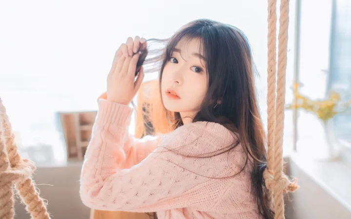 一名长发女子穿着粉色毛衣坐在室内秋千上。