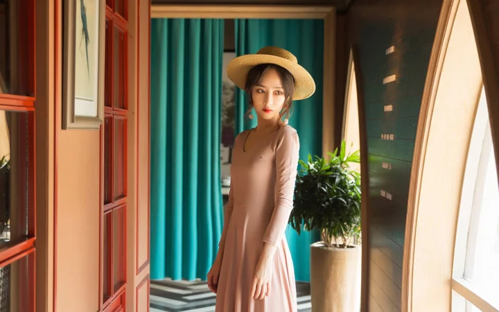 一位戴着宽檐帽、身穿长袖连衣裙的女性站在室内门边。