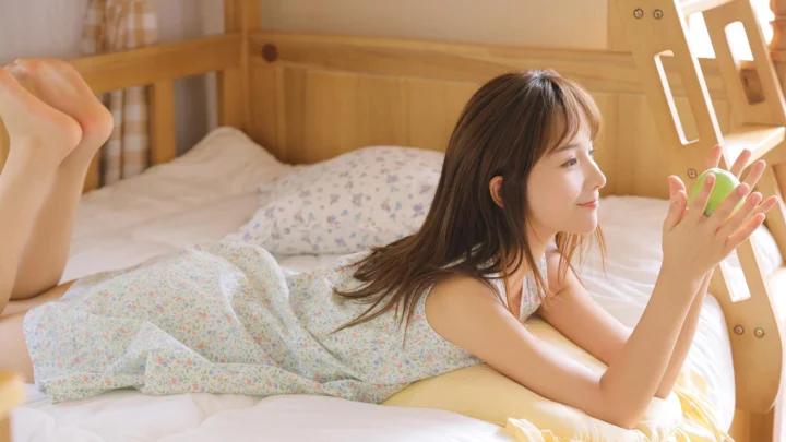 一名女子趴在阳光明亮的卧室床上，手中拿着一个绿色物体。
