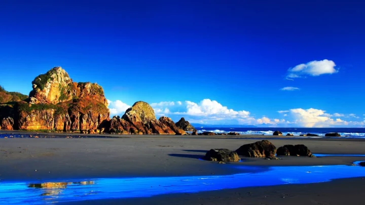 阳光照耀下的海滩延伸至远处的岩石海岸，前景映着湛蓝海水，天空中点缀着白云。