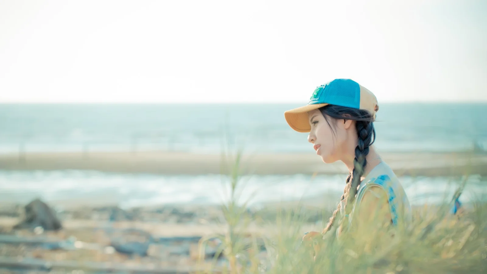 帽子をかぶった成人女性が海辺の草むらに横顔を見せて座り、背景に海と砂浜が広がっています。