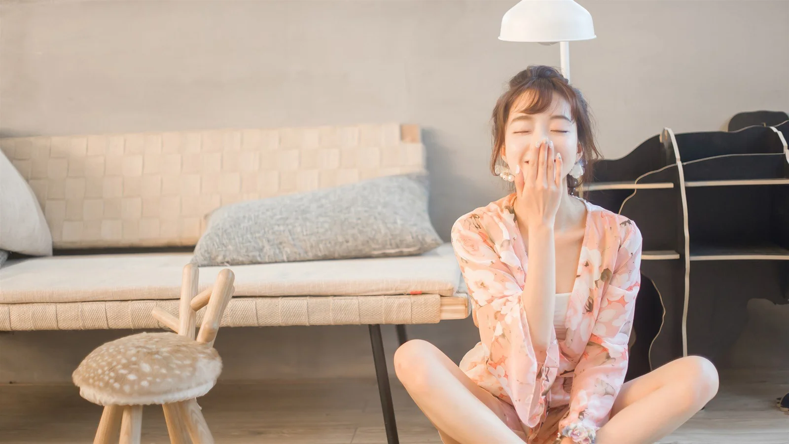 분홍색 꽃무늬 옷을 입은 성인 여성이 소파와 의자, 옷걸이가 있는 실내 바닥에 앉아 있습니다.