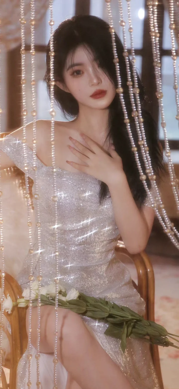 一名成年女性身穿闪亮的银色礼服，坐在珠帘前直视镜头。