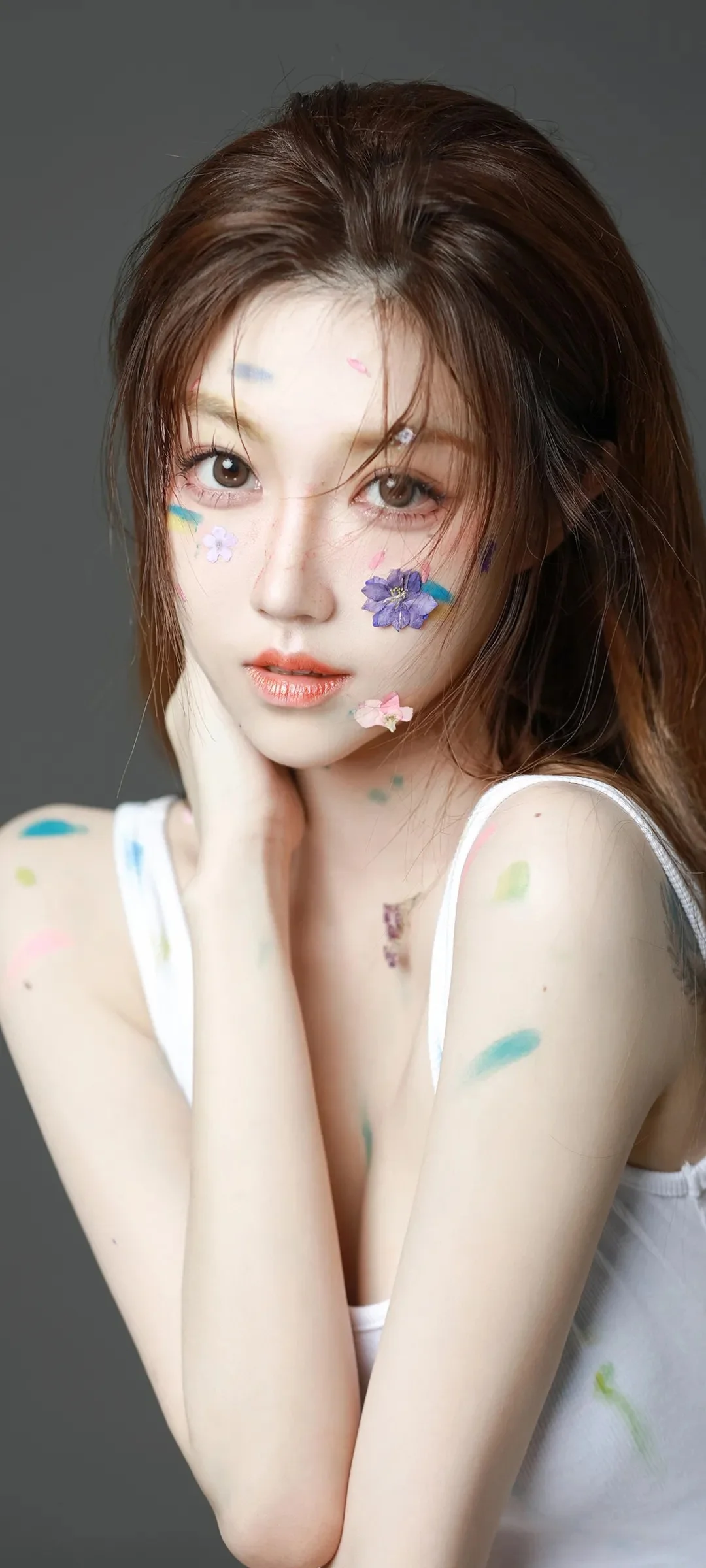 긴 머리의 여성이 흰색 민소매 상의를 입고 회색 배경 앞에서 얼굴의 꽃 장식을 보여준다.
