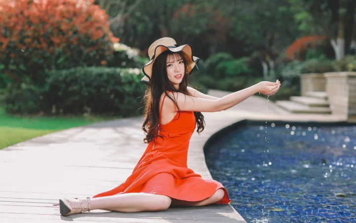 一名身穿红色连衣裙的女子坐在户外泳池边，身后是绿树和花园。