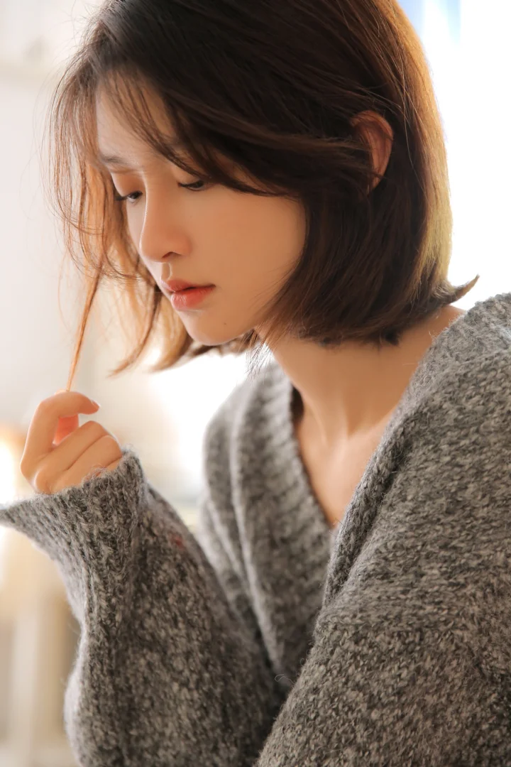 회색 니트를 입은 단발머리 여성이 밝은 배경 앞에서 옆모습으로 아래를 바라보고 있습니다.