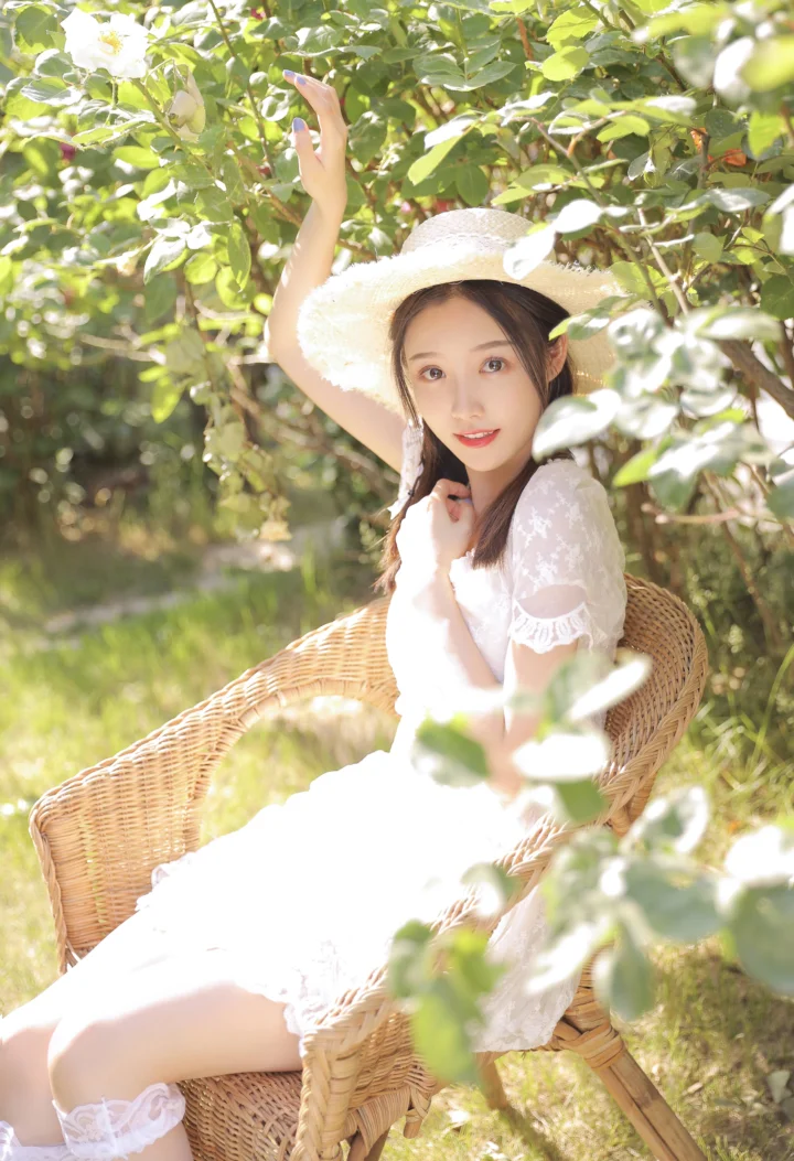一名戴草帽、穿白裙的女子坐在藤椅上，置身于阳光洒落的绿叶花园中。